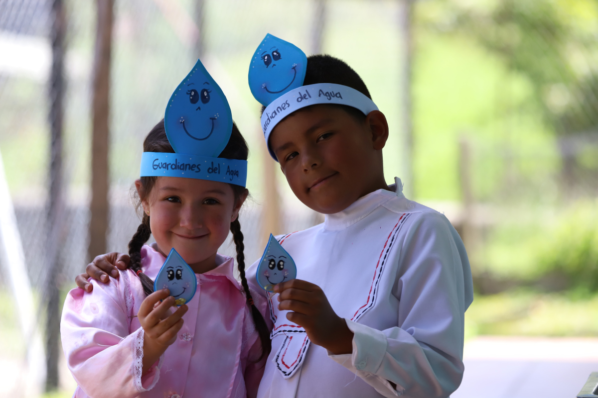 Dos niños sonríen a la cámara mientras usan diademas con el mensaje “Guardianes del Agua” y sostienen figuras con forma de gota durante una actividad educativa.