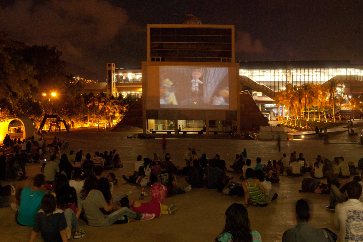 Personas reunidas en una plaza durante una proyección audiovisual al aire libre en la noche.