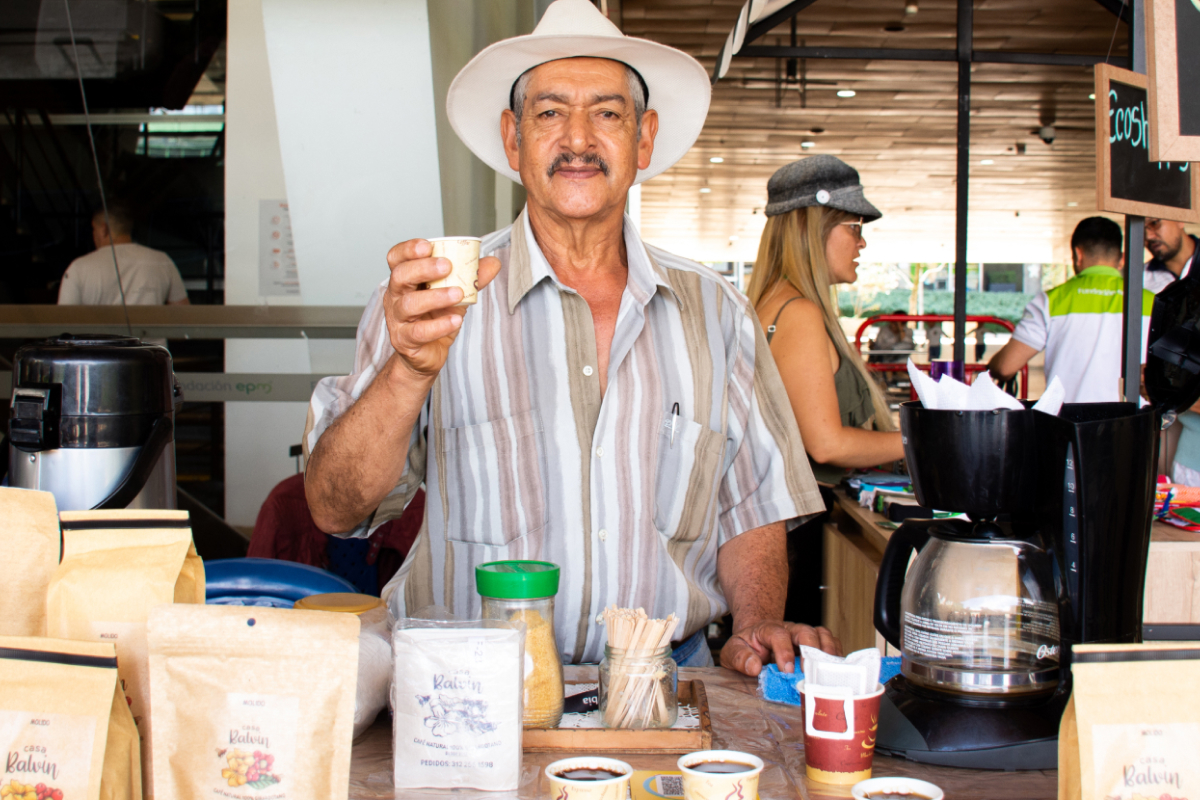 Hombre de pie en un puesto de café sosteniendo una taza pequeña, con bolsas de café y una cafetera sobre la mesa.