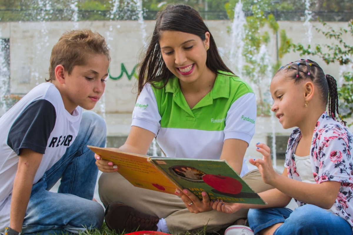 Educadora lee un libro a dos niños sentados al aire libre, en un espacio público con fuentes de agua al fondo.