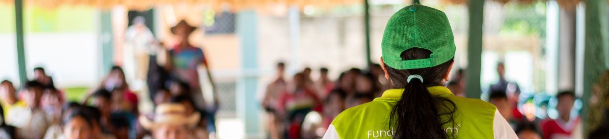 Persona con gorra y chaleco verde vista de espaldas, dirigiéndose a un grupo numeroso de personas reunidas en un espacio comunitario, en el contexto de una actividad educativa.