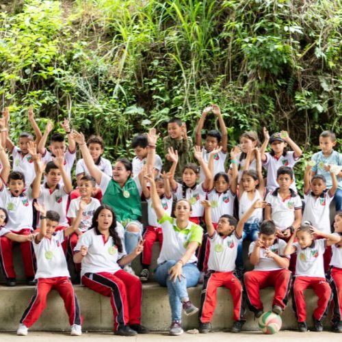 Grupo de niños y niñas con una persona adulta sentados al aire libre, levantando las manos en una actividad grupal.