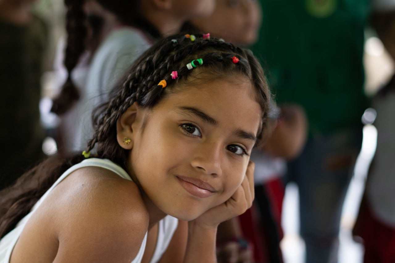 Niña con trenzas y cuentas de colores en el cabello sonríe mientras apoya el rostro en su mano, en un entorno escolar con otros niños al fondo.