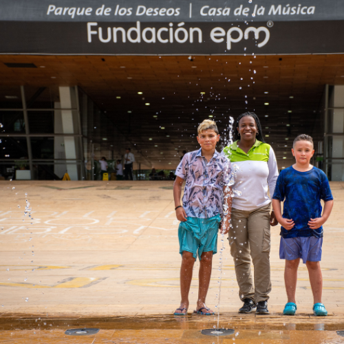 Mujer y dos niños de pie frente al edificio de la Casa de la Música de la Fundación EPM en el Parque de los Deseos, mientras chorros de agua de una fuente suben frente a ellos en un espacio público.