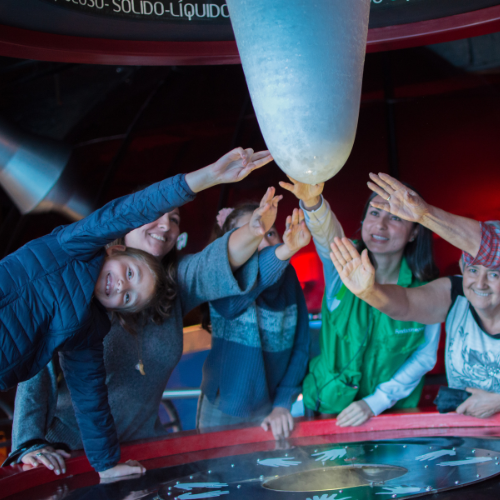 Grupo de niños y adultos interactúa con una exhibición científica, tocando un elemento suspendido dentro de un espacio educativo interactivo.
