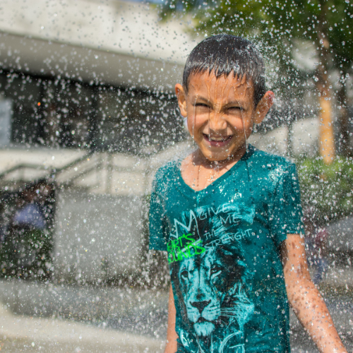 Niño jugando y sonriendo mientras el agua de una fuente lo salpica, representando alegría, bienestar y el disfrute del espacio público.