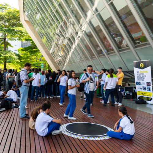Niños sentados alrededor de un juego interactivo mientras otras personas participan en una actividad educativa al aire libre frente a un edificio cultural.