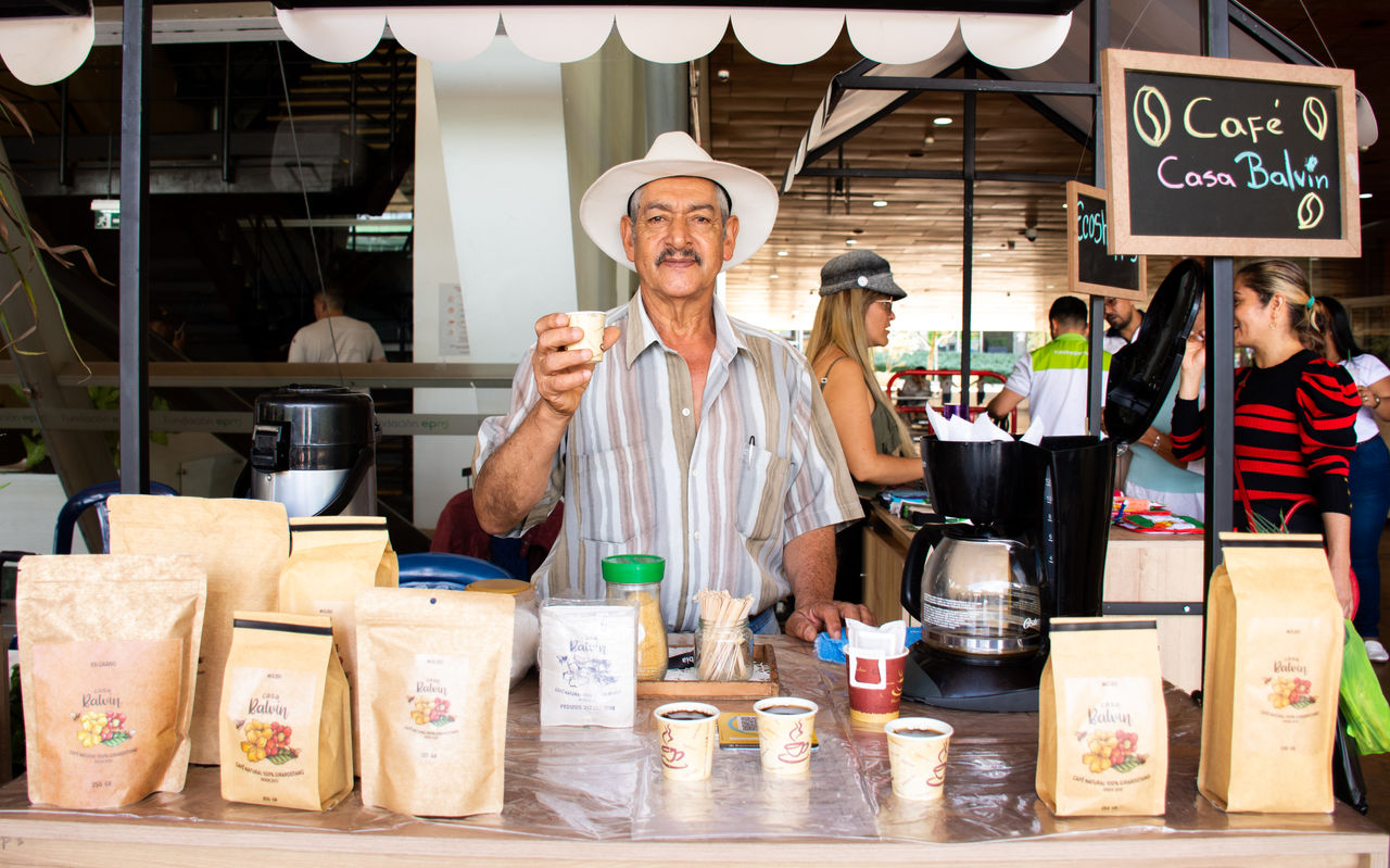 Hombre de pie en un puesto de café sosteniendo una taza pequeña, con bolsas de café y una cafetera sobre la mesa, y un letrero que dice “Café Casa Balvin”.
