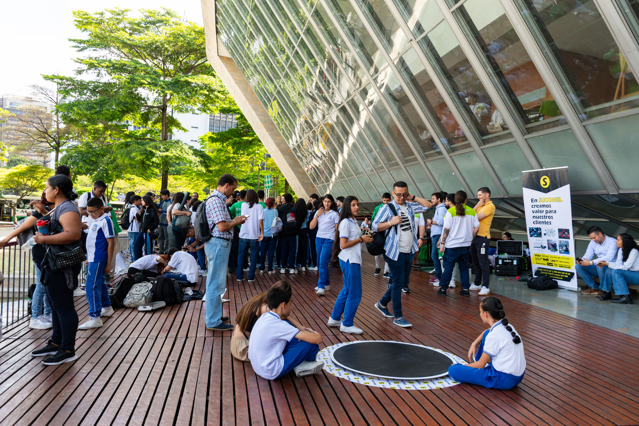 Niños sentados alrededor de un juego interactivo mientras otras personas participan en una actividad educativa al aire libre frente a un edificio cultural.