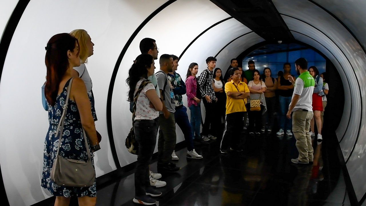 Grupo de personas de pie dentro de un pasillo curvo y cerrado, escuchando a un guía que habla al frente, con paredes claras y piso oscuro y brillante.