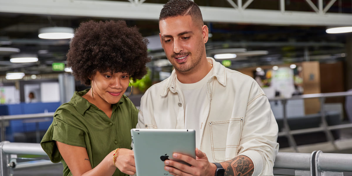 Dos personas, un hombre y una mujer, observando una tablet en un entorno interior, posiblemente un aeropuerto o estación.