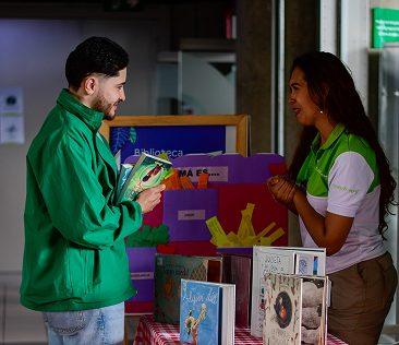 Dos personas conversan frente a un punto de biblioteca comunitaria, mientras revisan libros exhibidos sobre una mesa.