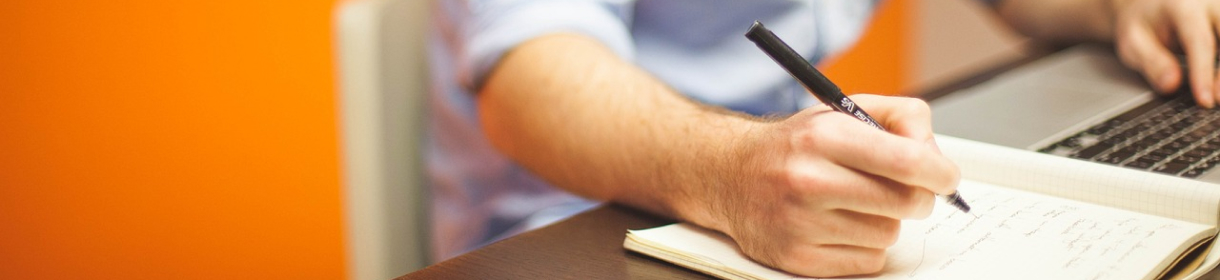 Persona escribiendo notas en una libreta junto a un ordenador portátil.